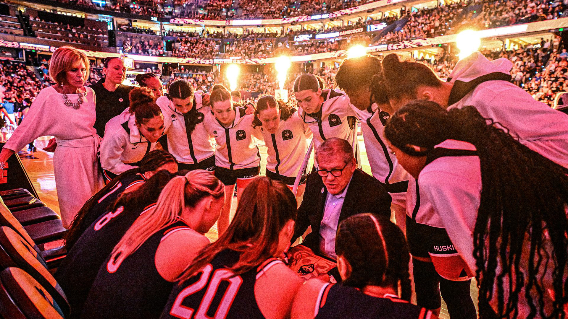 UConn women's basketball team players surrounding Coach Auriemma who is explaining a play in the middle of a game. 